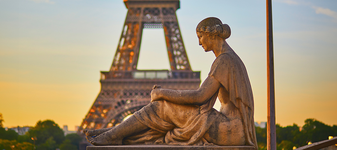 This image shows a classical stone sculpture of a seated female figure in the foreground, positioned in profile view with an elegant hairstyle and draped clothing. In the background, the Eiffel Tower can be seen during what appears to be sunset or sunrise, with the sky displaying warm orange and blue hues. The sculpture is likely located at the Trocadéro Gardens or another Parisian location that offers views of the Eiffel Tower. The juxtaposition of the historical sculpture against Paris's iconic landmark creates a quintessential scene representing French art and culture.