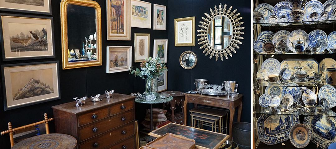 This image shows the interior of an antique shop or vintage decor boutique with several distinct elements. The space features a dark navy blue wall adorned with a gallery-style arrangement of framed artwork and prints, including what appears to be nature and landscape scenes. A decorative sunburst mirror with a gold finish serves as a focal point on the wall. The room contains several pieces of brown wooden furniture including a chest of drawers, small tables, and what looks like nesting tables. There are decorative silver objects displayed, including small animal figurines on one of the dressers and some silver serving pieces. A vase with greenery sits on a round table in the center. On the right side of the image is a prominent display of blue and white porcelain dishware - plates, bowls, and serving pieces arranged on shelves. The collection appears to be in the traditional blue and white willow or delft style. The overall aesthetic suggests a curated collection of antiques and collectibles in a sophisticated, traditional setting.