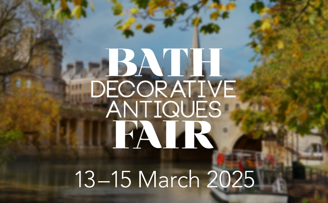 This promotional image advertises the "Bath Decorative Antiques Fair" taking place from March 13-15, 2025. The text is prominently displayed in elegant white typography against a softly blurred background featuring the historic city of Bath, England. The scene captures autumn trees with yellow and green leaves framing the view, with the city's distinctive honey-colored Georgian architecture visible in the background. A glimpse of water (likely the River Avon) appears at the bottom of the image, suggesting Bath's picturesque riverside setting. The overall aesthetic combines sophistication with a seasonal warmth that complements the antiques theme of the event.