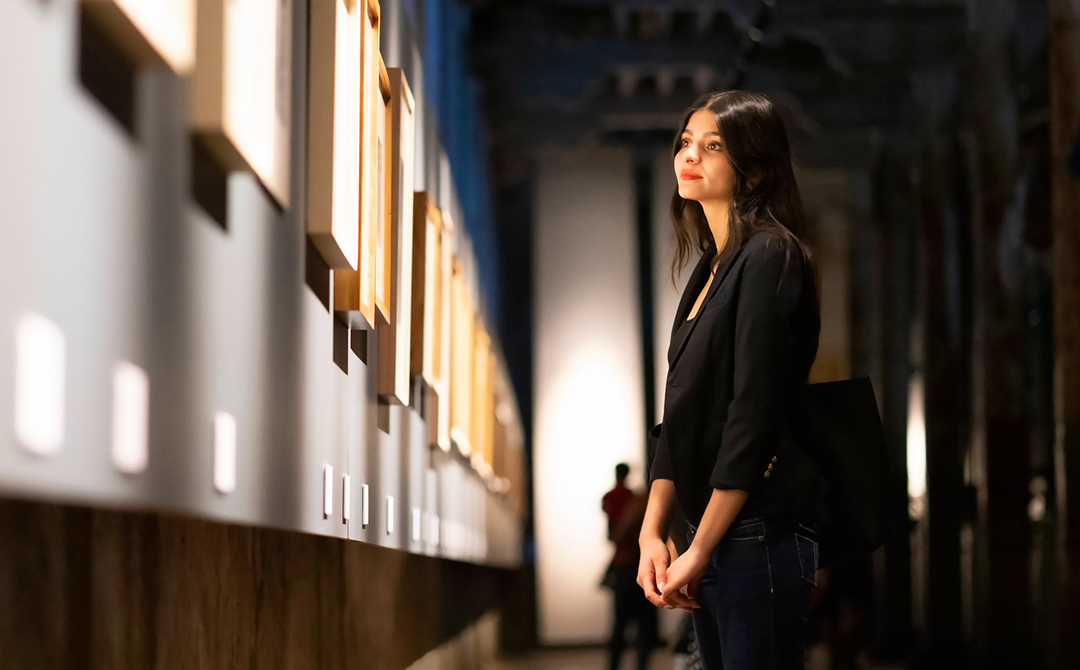 Young woman in a black blazer thoughtfully viewing framed artwork in a softly lit gallery, capturing the spirit of discovery central to antiques fairs.