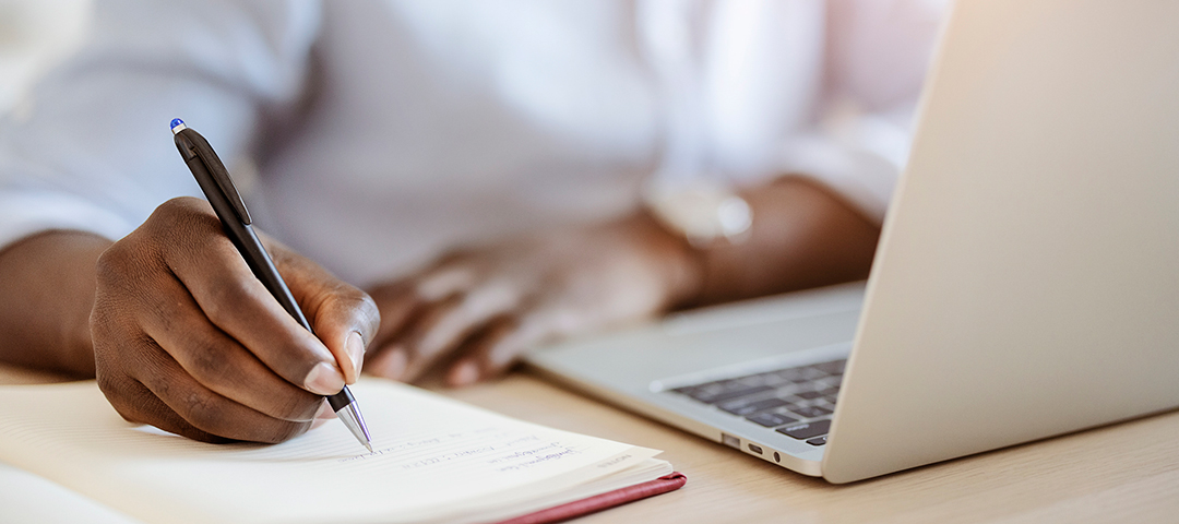 Dealer writing inventory notes beside a laptop, representing the shift from handwritten records to digital cataloging for antique sellers.