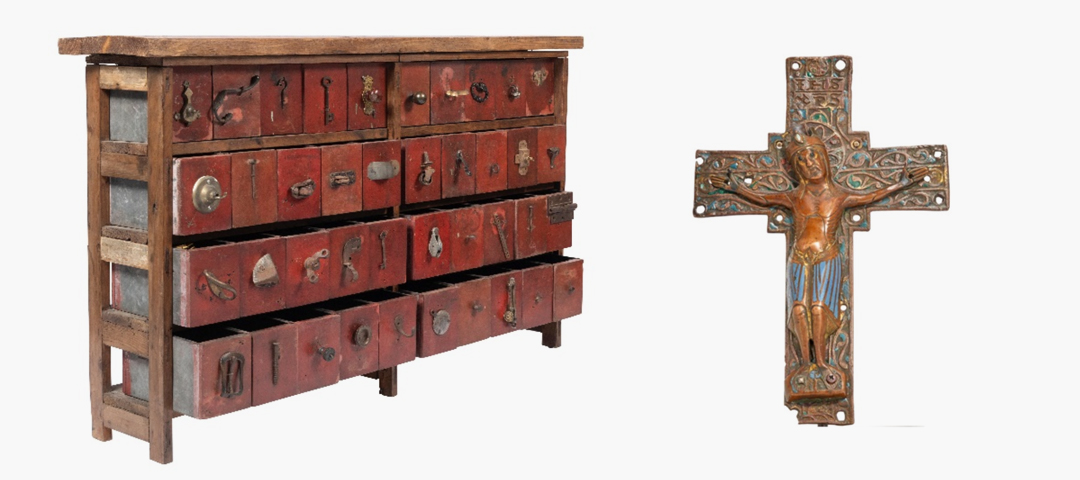 Antique wooden chest with red-painted drawers and varied metal pulls displayed beside a decorative painted crucifix, highlighting utilitarian furniture and religious folk art at The Decorative Fair.