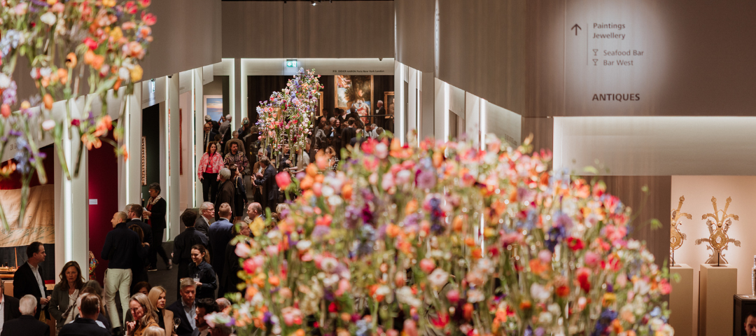 Crowded interior corridor at a major European antiques fair with gallery booths and floral installations, illustrating institutional-level presentation and international attendance.