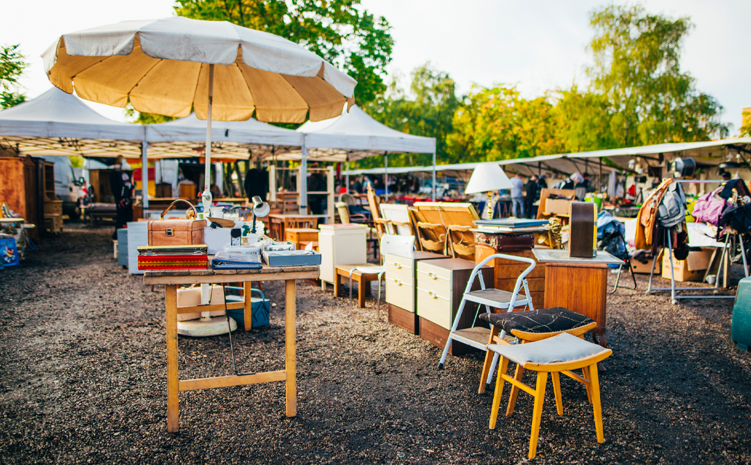 Open-air flea market with vintage furniture, lamps, and small antiques displayed under vendor tents as buyers browse for unique pieces.