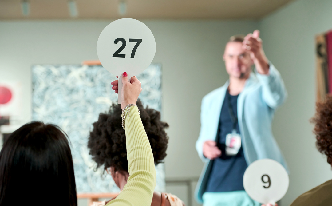 Interior designer raises paddle number 27 during a live auction as bidding progresses quickly, illustrating the decisive timing required to secure one-of-a-kind vintage and antique pieces.