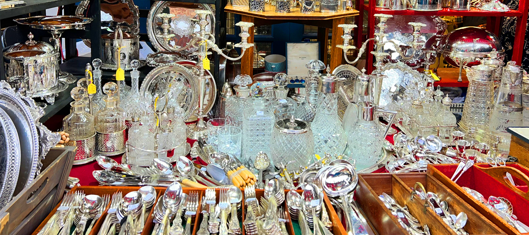 Table filled with antique silverware, glass decanters, serving trays, and decorative objects displayed at a flea market stall.