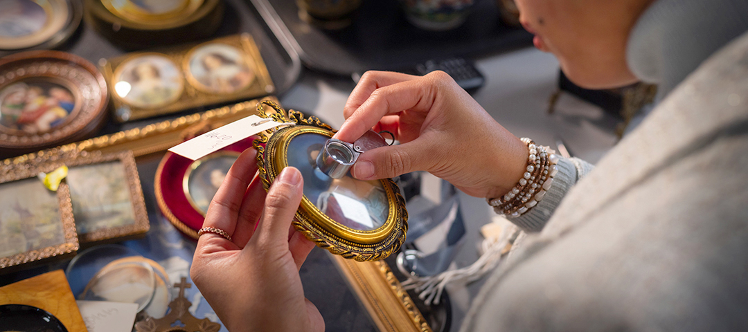 A collector examines a framed miniature portrait with a magnifying loupe at a design fair, highlighting close study and connoisseurship in the antiques market.