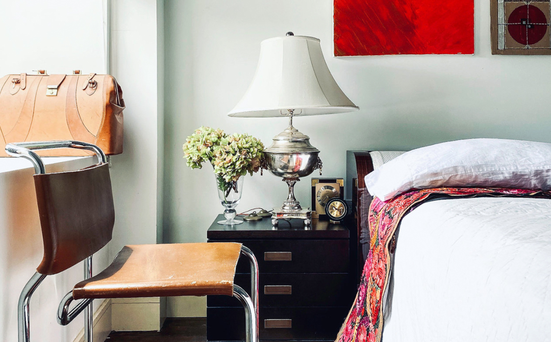 A layered bedside vignette featuring a vintage leather chair, antique silver lamp, and framed artwork, combining modern bedding with collected, one-of-a-kind pieces.
