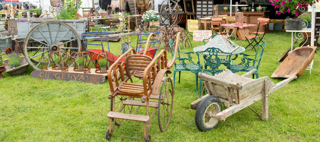 Outdoor display of vintage garden antiques and architectural salvage, including wooden carts, iron seating, and decorative objects arranged on grass