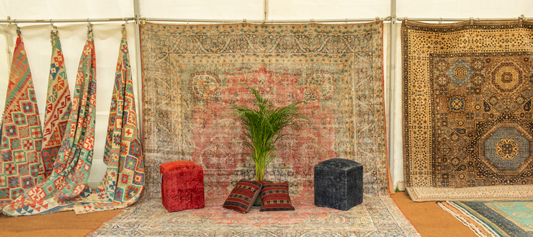 Layered display of vintage and antique rugs with upholstered stools and textiles, showing color, pattern, and material variation