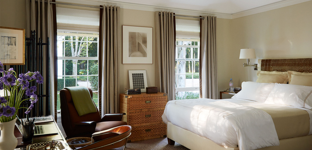 A serene bedroom by David Kleinberg layered with woven textures, tailored drapery, vintage furnishings, and understated neutral tones inspired by the quiet luxury aesthetic of the 1990s.