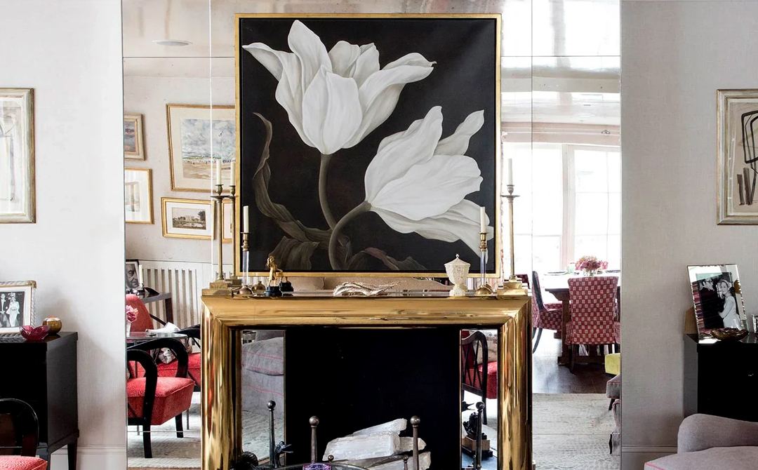 Nina Campbell interior with a polished brass mantel, a large black-and-white floral painting, and salon-style framed art layered across mirrored walls.