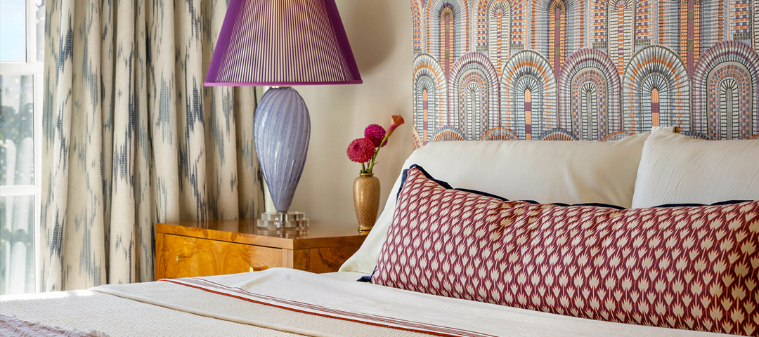 Bedroom designed by Michelle Workman Interiors featuring an Art Deco arched-motif headboard, a burl-wood nightstand, and a pleated violet lamp.