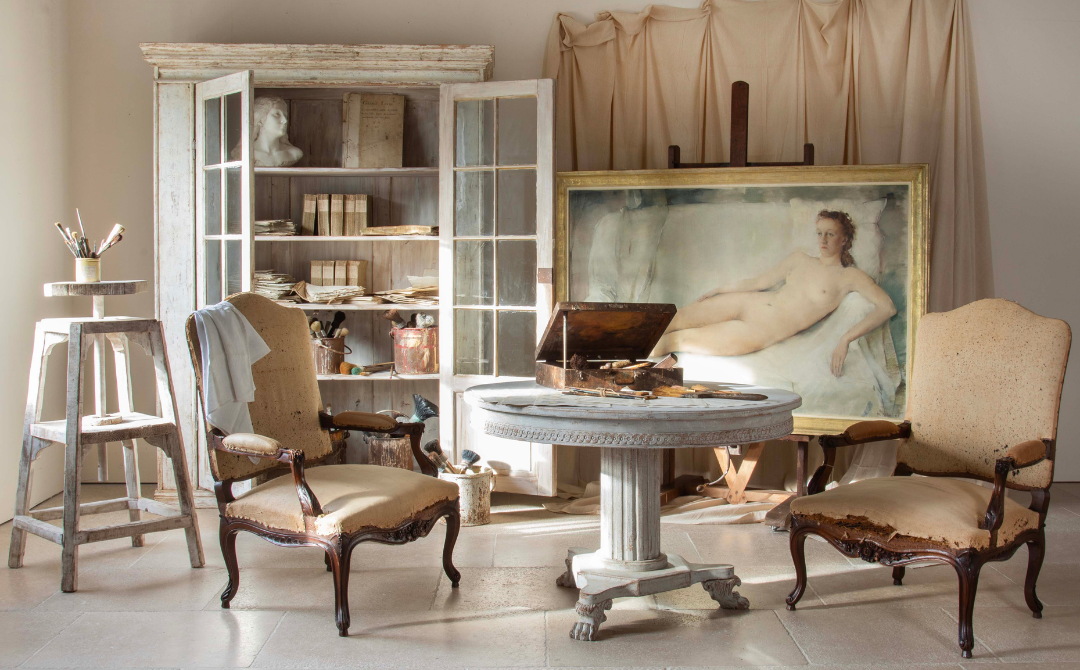 A pair of Louis XV fauteuils stripped to frame and burlap, their cabriole legs and carved crest rails exposed, in an artist's studio beside a painted French vitrine and pedestal table.