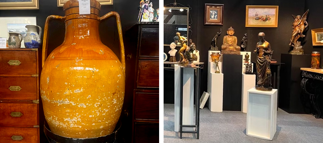 Large antique glazed terracotta urn beside a campaign chest, and a display of bronze figural statues on pedestals at an antiques fair