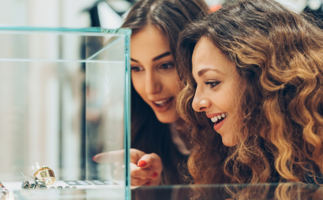 Two women browsing antique jewelry in a glass display case, pointing out a piece and smiling — capturing the hands-on discovery that's drawing a new generation to antique