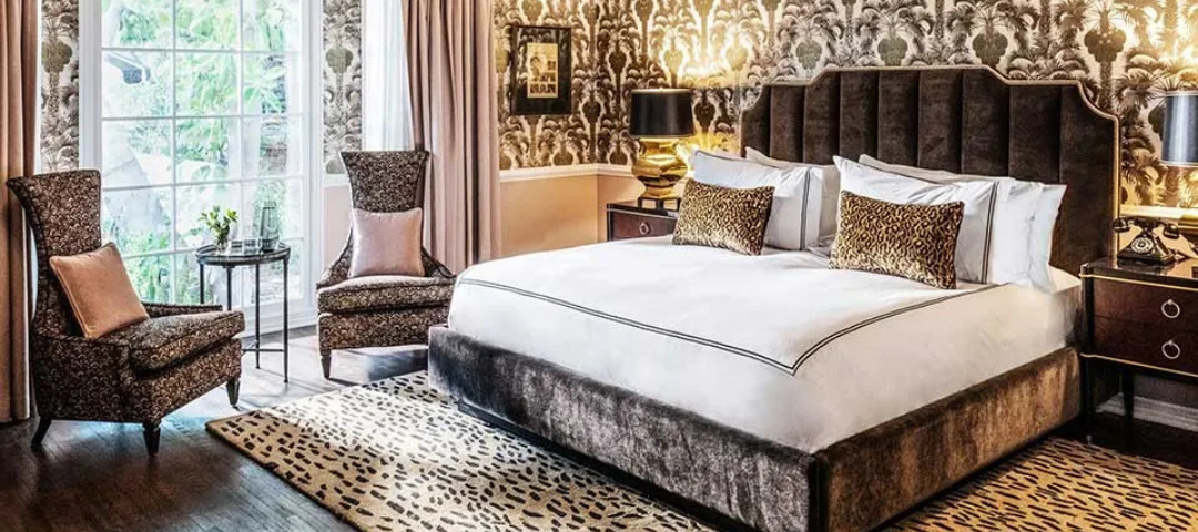 Art Deco-inspired bedroom with a channel-tufted velvet bed, leopard-print rug, gold table lamps, and palm-motif wallpaper, pairing bold pattern with disciplined symmetry created by Martyn Lawrence Bullard.