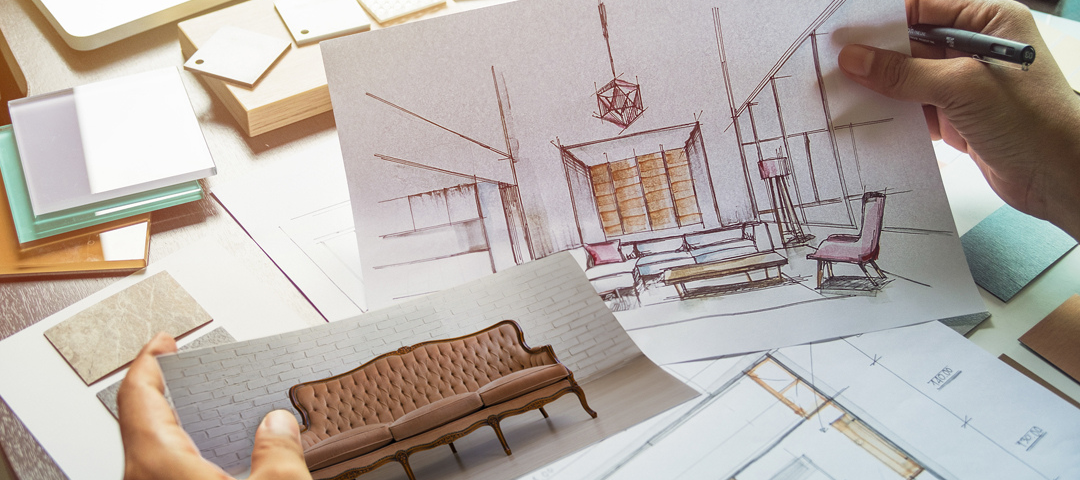 A designer's desk with an interior sketch, material and stone swatches, and a hand holding a photo of a tufted antique sofa against a floor plan.