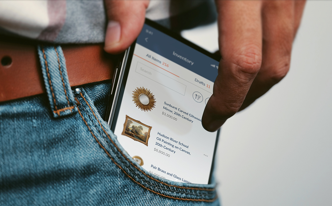Smartphone displaying the Ronati Mobile App inventory list, tucked into a jean pocket, showing antique items with prices.