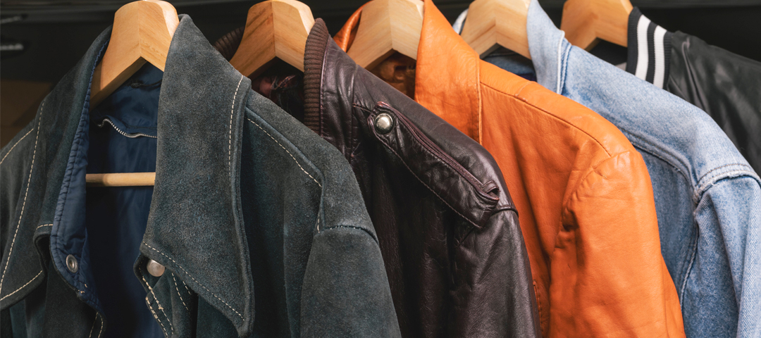 A rack of vintage leather and denim jackets in varied colors, hanging on wooden hangers.