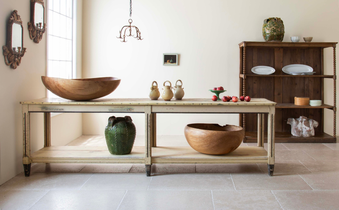 A curated room of one-of-a-kind antiques inventory: a long painted work table, carved wooden bowls, green glazed jars, and a bobbin-turned open shelf