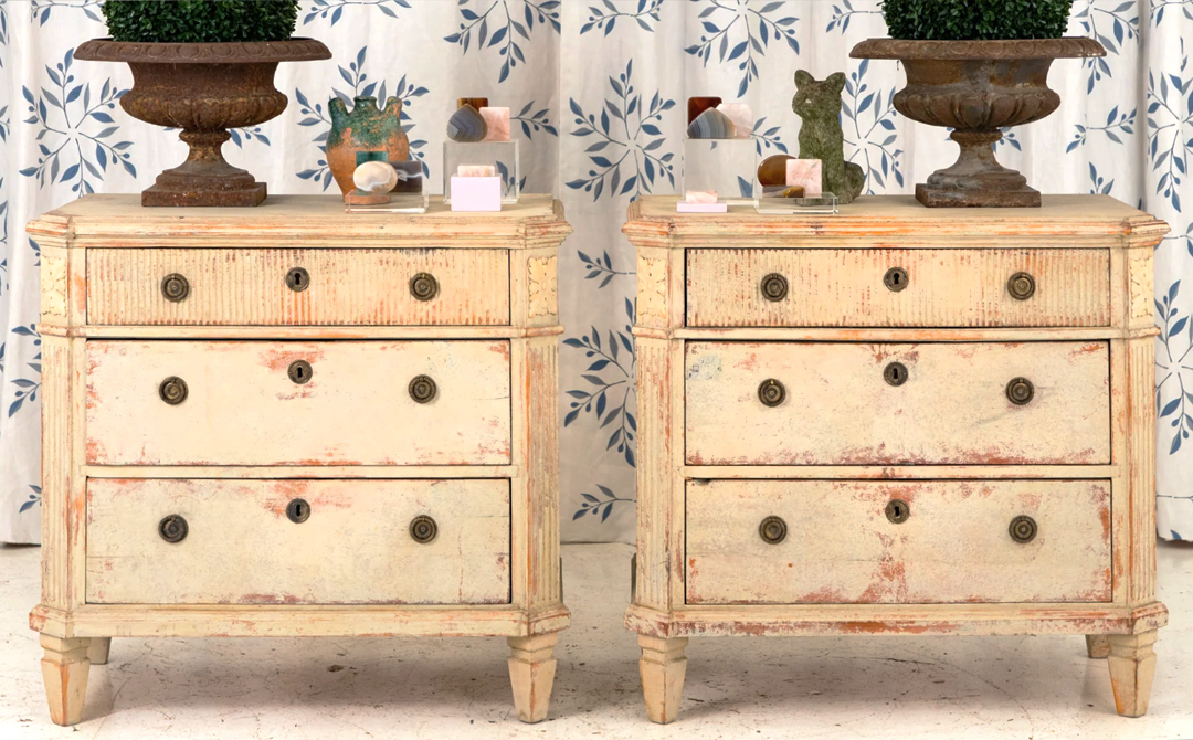 Pair of Swedish Gustavian chests of drawers with worn painted finish, flanking urns and small objects.
