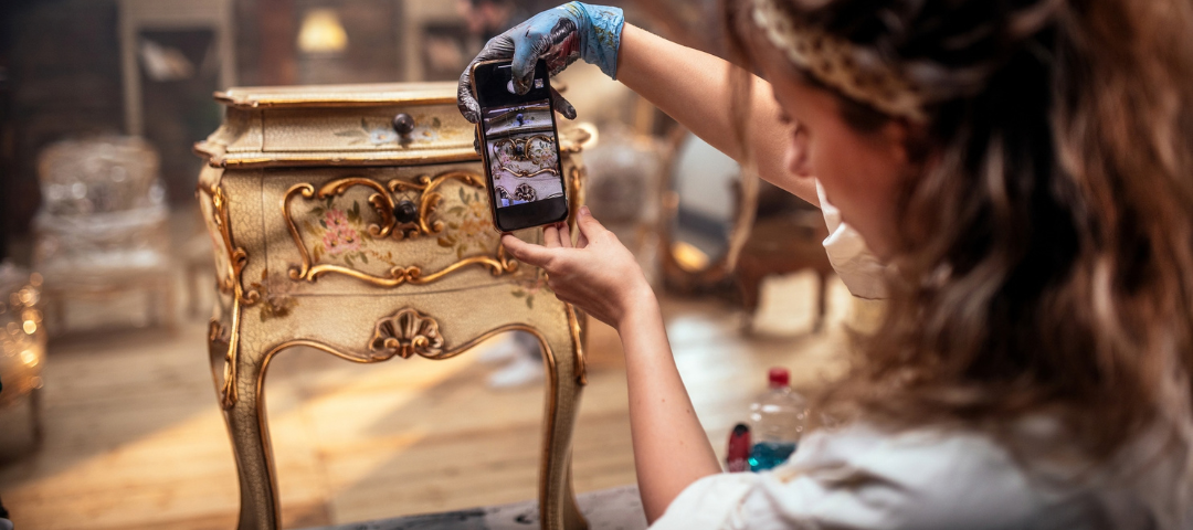 A woman in work gloves photographs a painted and gilded bedside cabinet with her phone, documenting the piece at a fair