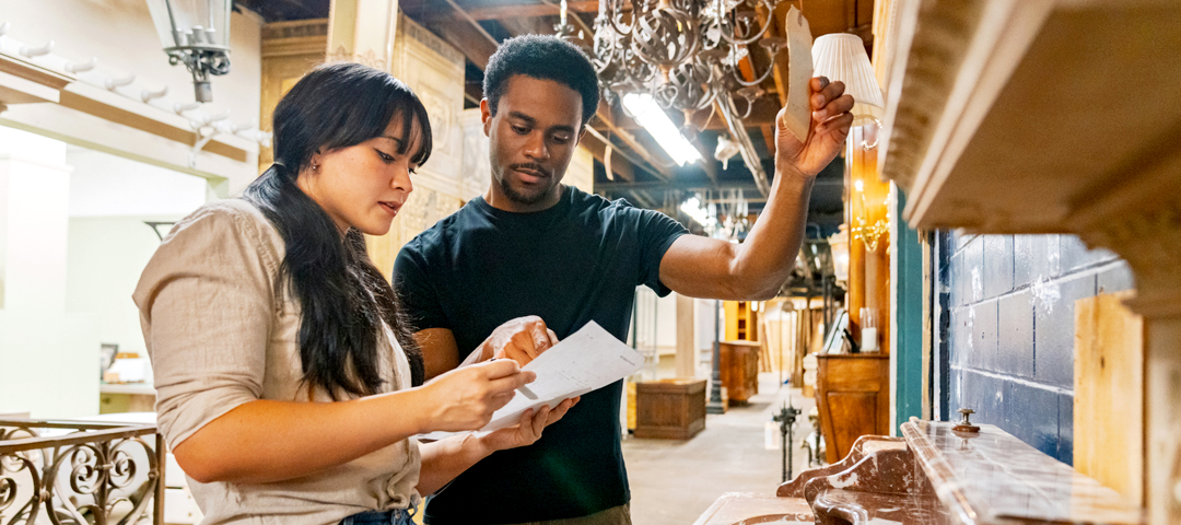 Two dealers review paperwork beside a tagged chandelier in an architectural salvage warehouse filled with antique mantels and lighting