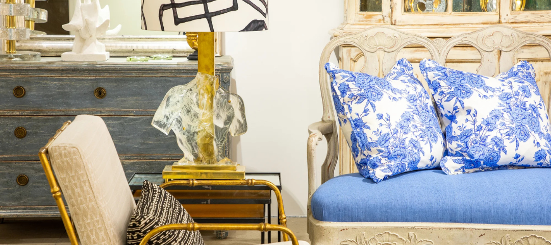 Vignette pairing a painted grey Swedish chest and mirrored sculptural lamp with a carved cream settee dressed in blue floral pillows.