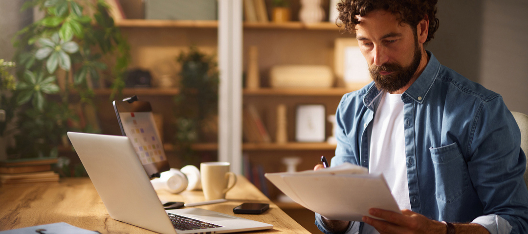 A dealer reviews paperwork with a pen in hand at his desk, a laptop and tablet beside him, tallying the costs connected to a piece of inventory