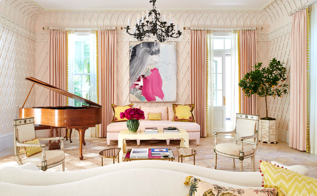Pink living room with trellis-paneled walls, where a pair of painted neoclassical armchairs and a grand piano sit beneath a bold abstract painting and an iron chandelier.