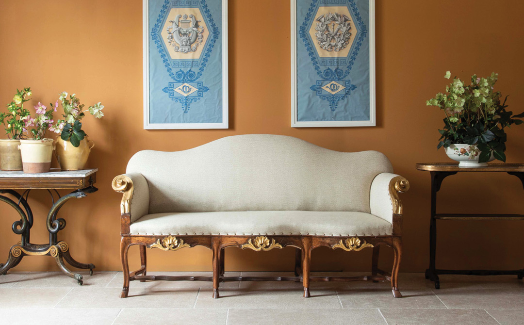 Antique carved and gilded settee in cream upholstery against an ochre wall, flanked by side tables with potted flowers.