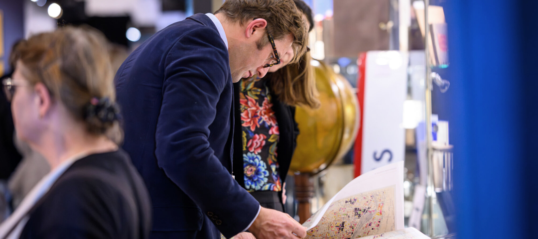 Visitors at an antiques fair lean in to examine an antique map at a dealer's booth, with a globe behind them.