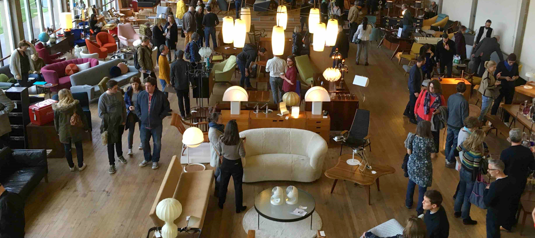 Overhead view of buyers browsing vintage furniture and lighting on a crowded antiques fair floor.