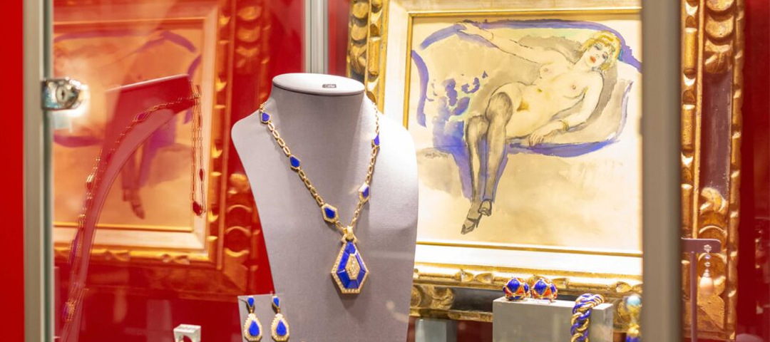 Gold necklace and earrings set with deep blue stones on a display bust, in front of a gilt-framed watercolor of a reclining figure