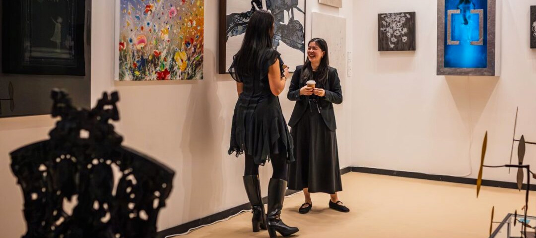 Two visitors in conversation in a contemporary art booth hung with abstract and floral paintings, with a dark sculptural chair in the foreground